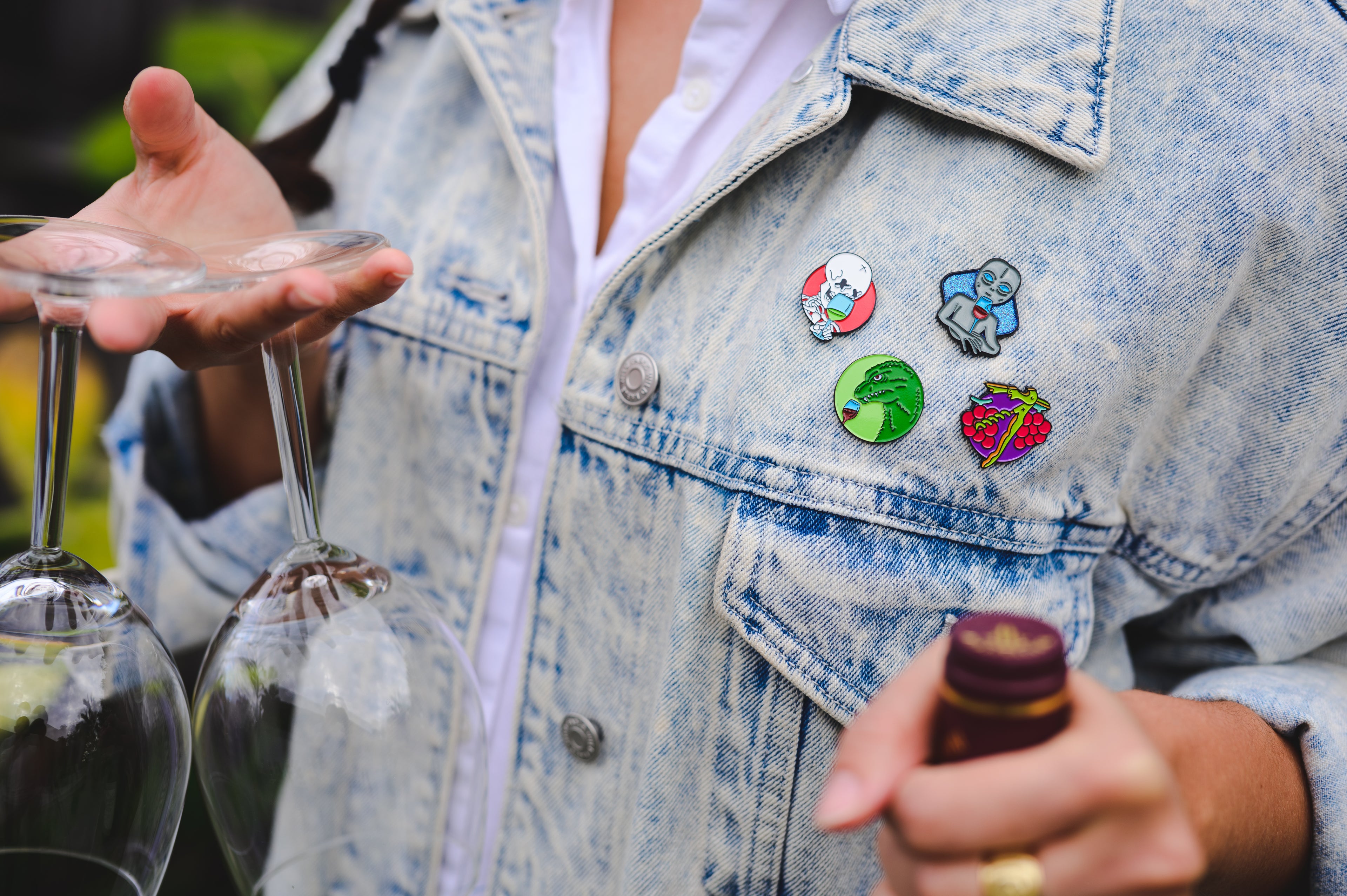 A person wearing a jean jacket with four shitty wine memes wine enamel pins on.  Winezilla, wine skull pin, alien wine pin, and a corkscrew pin. There are also holding two wine glasses and a wine bottle.